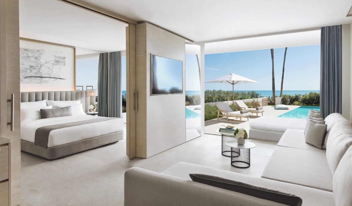 Modern suite at Ikos Andalusia with bed, lounge area, and ocean-view terrace with pool, loungers, and white umbrella.