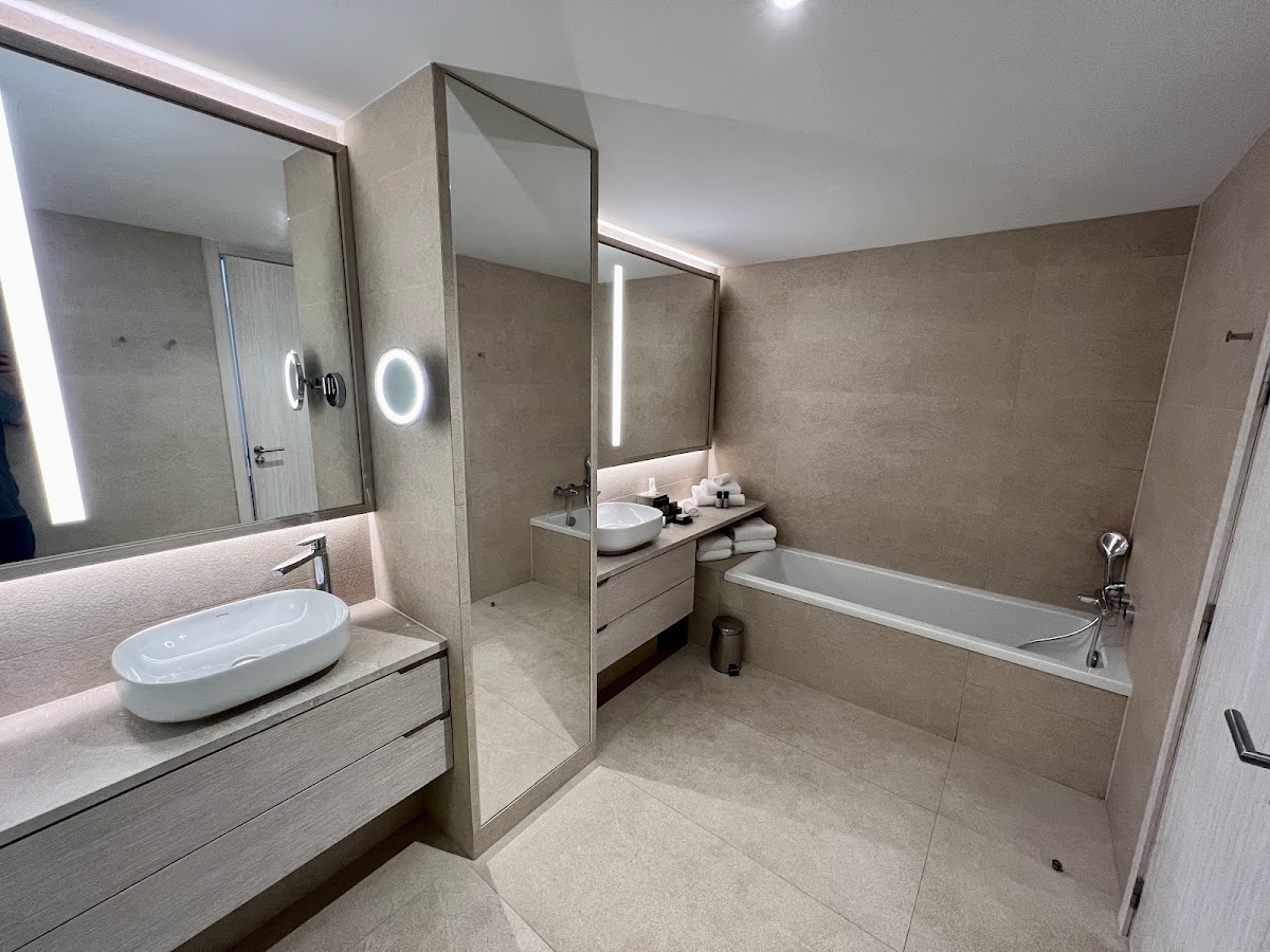 Modern bathroom at Ikos Andalusia with dual vanities, illuminated mirrors, walk-in shower, and soaking tub in neutral stone tones.