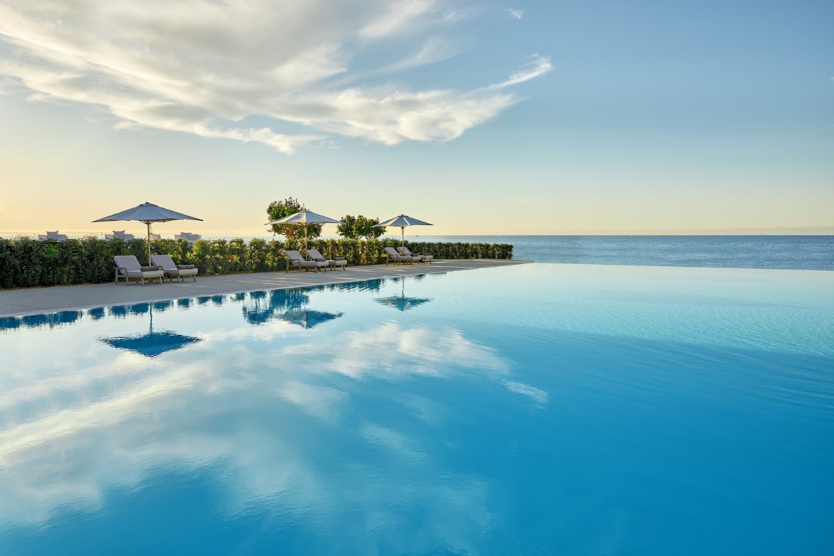 Infinity pool overlooking the ocean at Ikos Andalusia with loungers, blue umbrellas, and green landscaping along the shore.