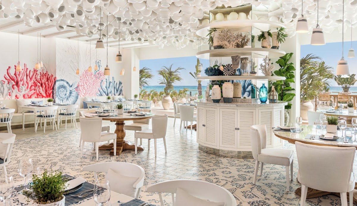 Bright coastal restaurant with white furniture, coral and blue artwork, open shelving with decorative objects, and ocean views through large windows.