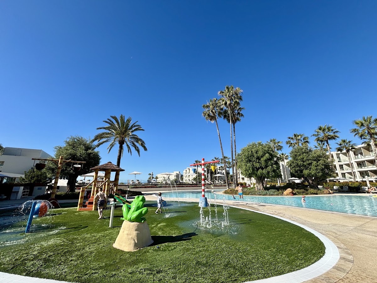 Children's play area with green lawn, colorful sculptures, and pools at Ikos Andalusia under clear blue sky.