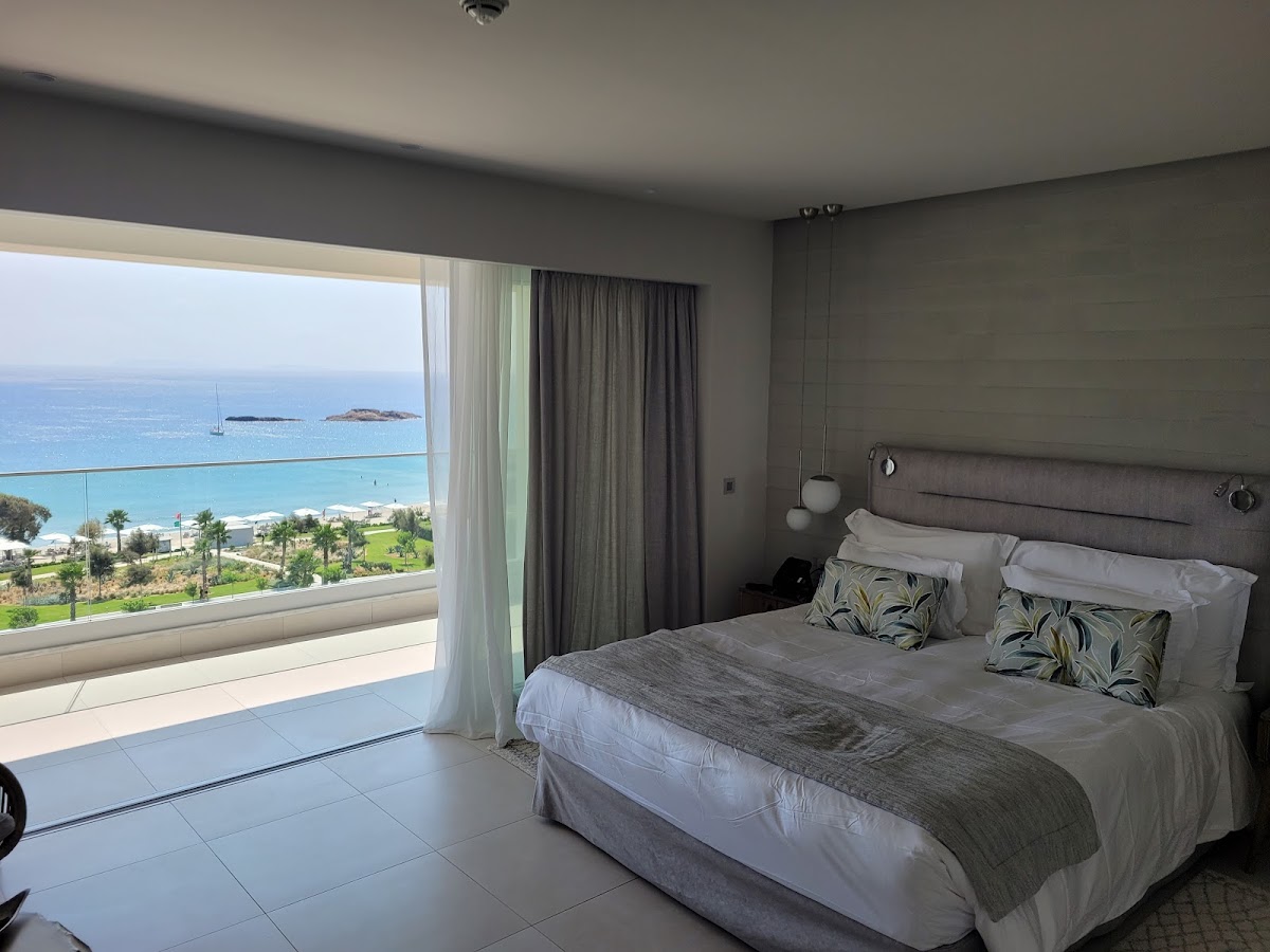 Bedroom with gray upholstered bed facing floor-to-ceiling windows overlooking Ikos Aria's beach, turquoise sea, and distant islands.