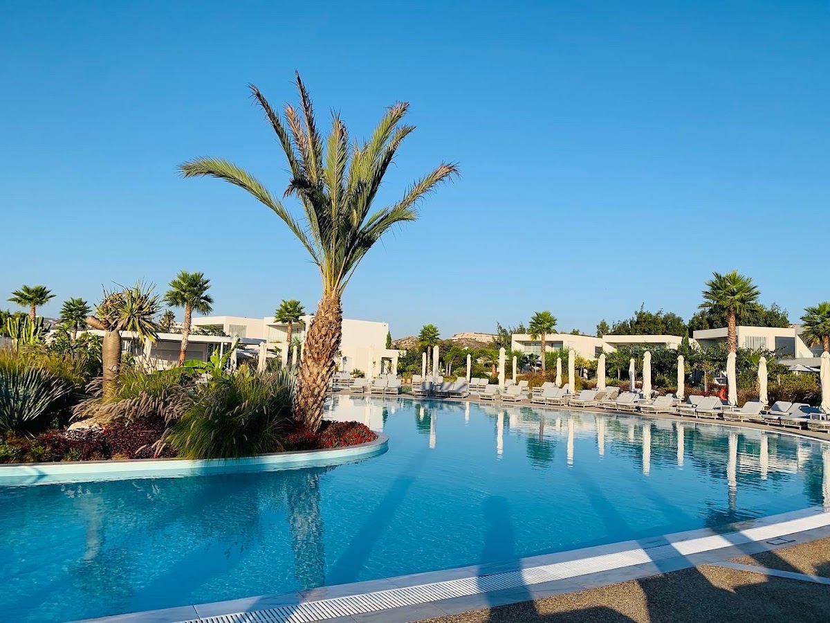 Large resort pool with turquoise water, white columns, palm trees, and cream-colored buildings under clear blue sky at Ikos Aria.