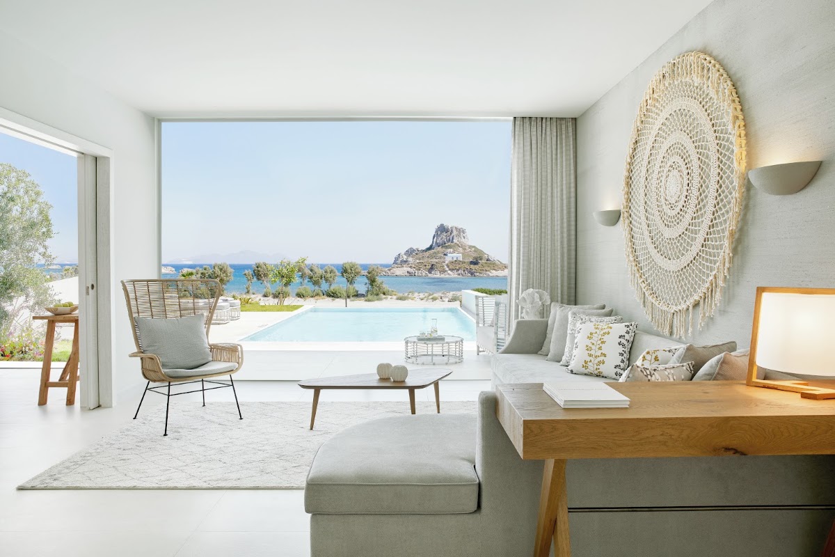 Modern suite at Ikos Aria with sea view of rocky island, infinity pool, woven wall decor, and furnished terrace overlooking turquoise waters.