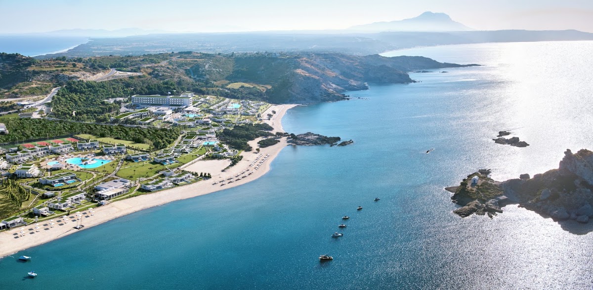 Aerial view of Ikos Aria resort on a turquoise bay with sandy beach, green hillsides, and rocky islands in the distance.