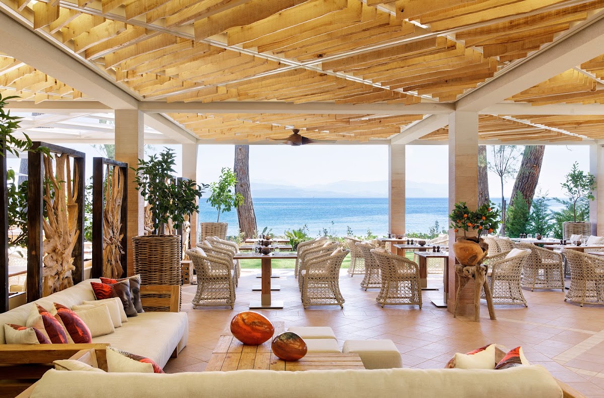 Beachfront dining and lounge area at Ikos Aria with wooden ceiling beams, woven chairs, cream seating, and ocean view.