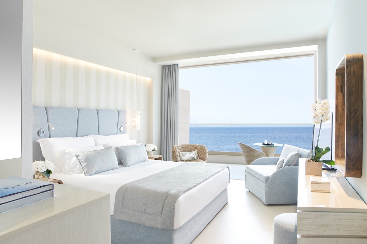 Bright beachfront bedroom at Ikos Dassia with white bedding, blue headboard, and expansive sea views through floor-to-ceiling windows.