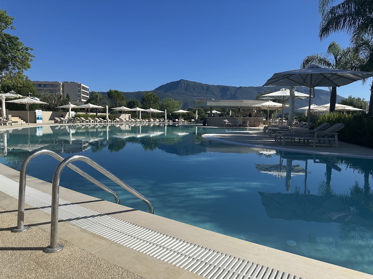 Ikos Dassia pool with metal entry ladder, sunbeds, umbrellas, and mountains in the background under clear blue sky.