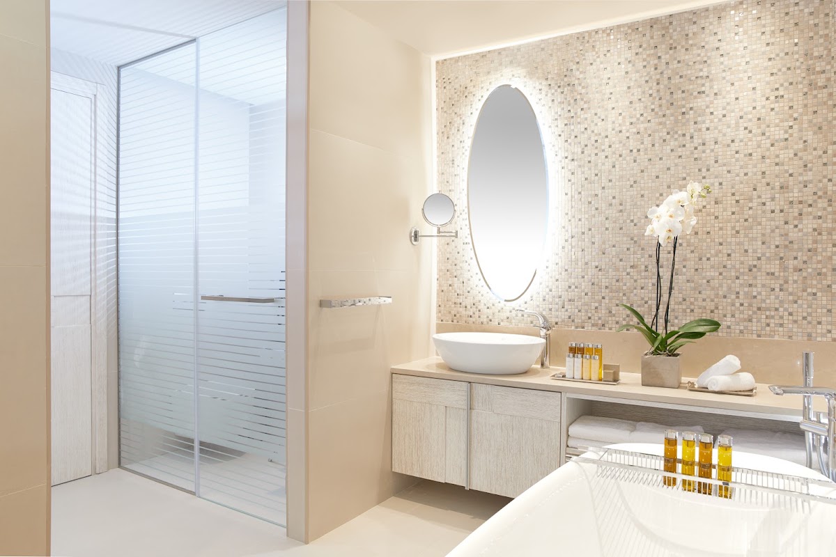 Resort bathroom with mosaic tile wall, oval lit mirror, white vessel sink, glass shower enclosure, and orchid plant on vanity.
