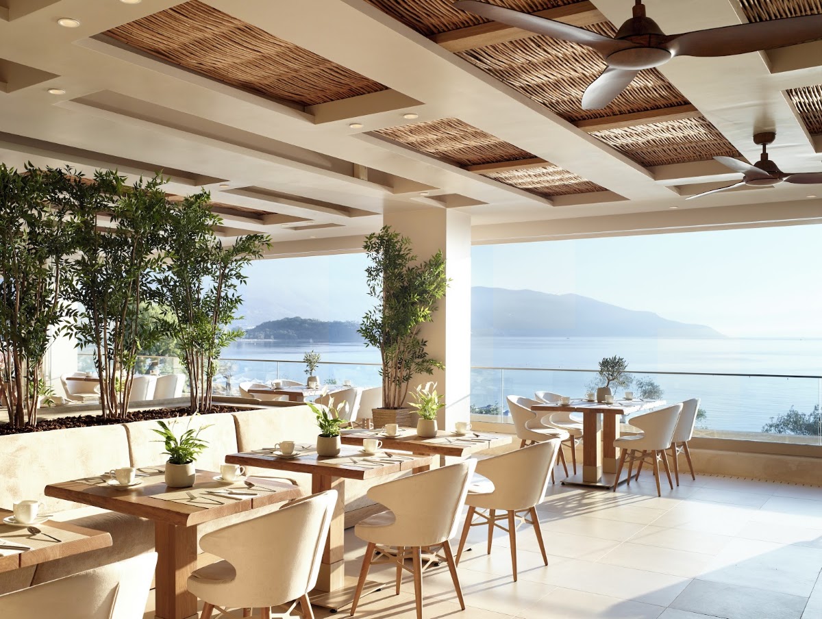 Coastal dining area at Ikos Dassia with wooden tables, cream chairs, and woven ceiling details overlooking a calm bay and distant mountains.