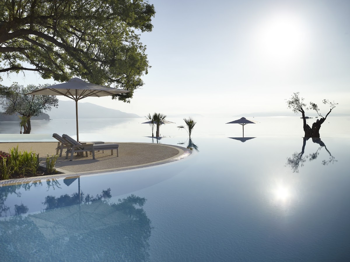 Infinity pool with loungers and umbrellas overlooking a calm bay, palm trees and gnarled olive tree visible, mountains across the water.