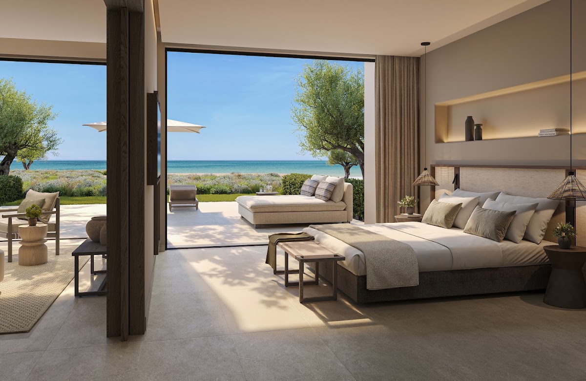 Beachfront suite at Ikos Kissamos with bed, patio doors opening to terrace with lounge seating, and Mediterranean Sea view beyond.