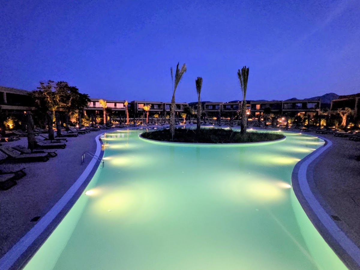 Illuminated lagoon-style pool at Ikos Kissamos glows turquoise at dusk, surrounded by lounge chairs and palm trees with lit resort buildings beyond.