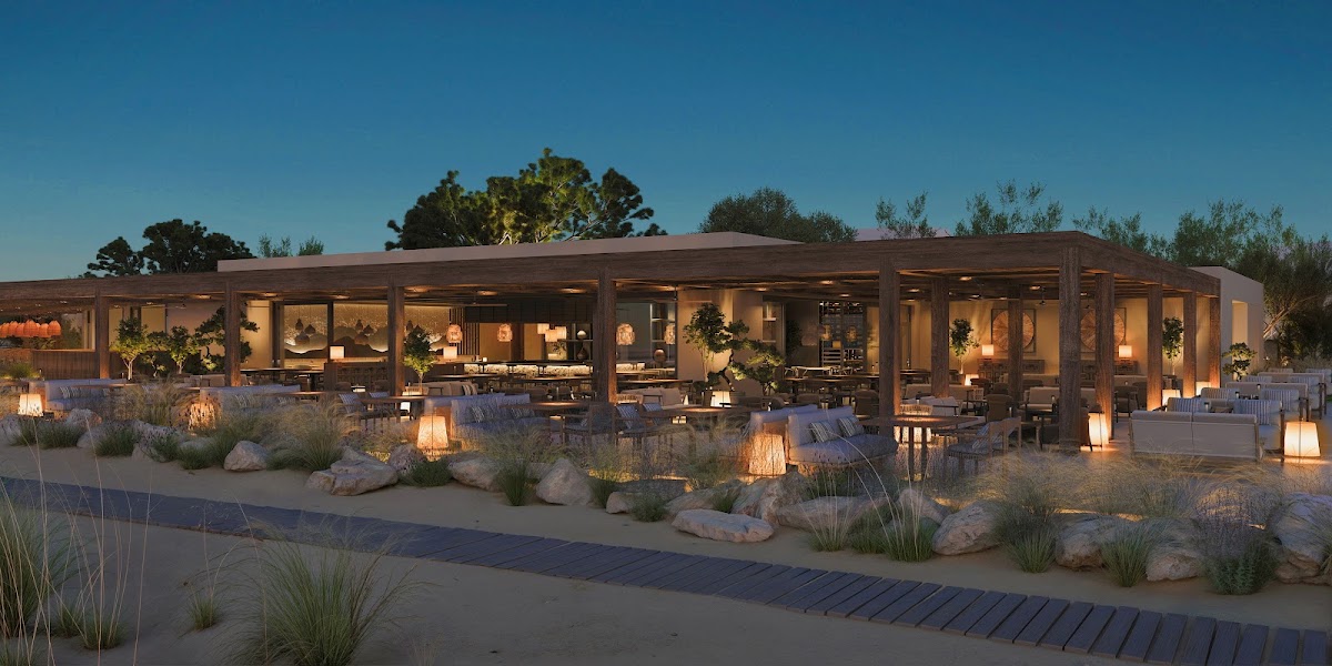 Beachfront dining pavilion at Ikos Kissamos lit by lanterns at dusk, with sand, coastal grasses, and rocks in foreground.