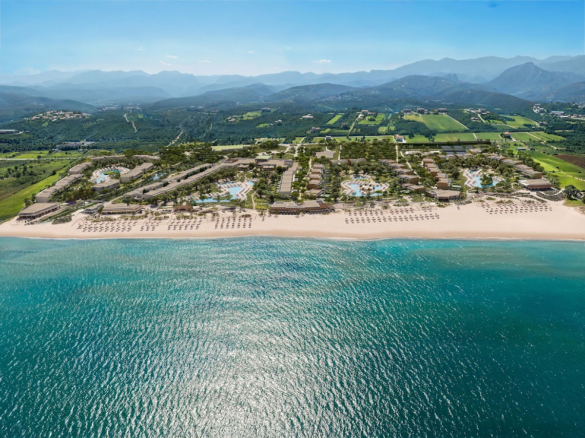Aerial view of Ikos Kissamos resort on a sandy beach with turquoise water, multiple pools, and buildings nestled among green landscaping, with mountains rising beyond the valley.