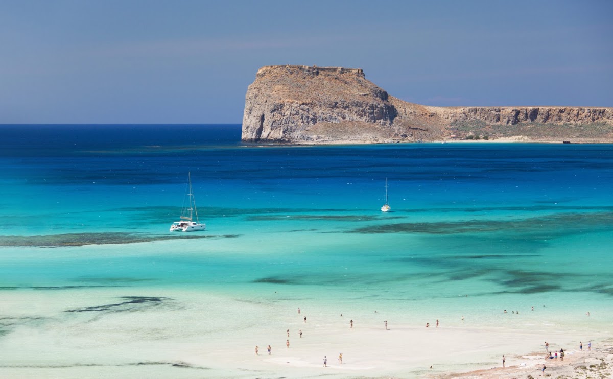 Swimmers and boats in turquoise waters below a towering rocky headland at Ikos Kissamos.