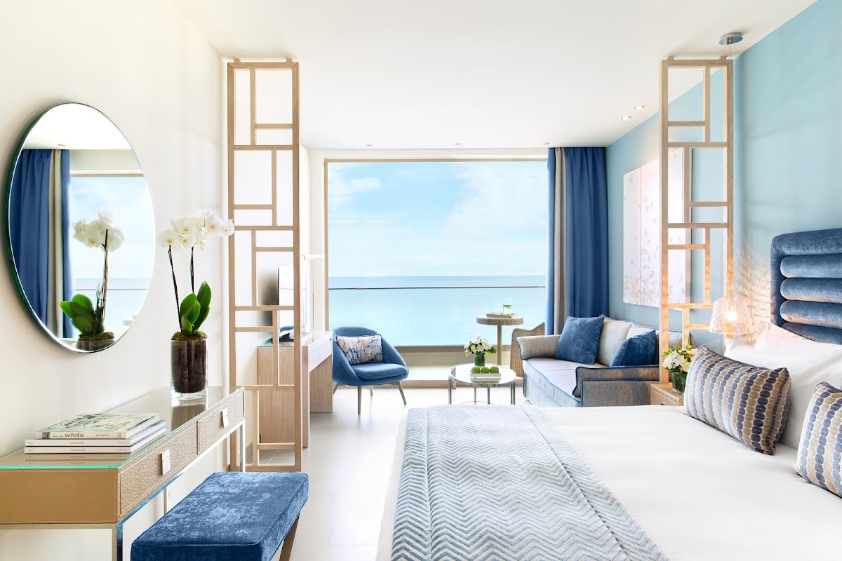 Oceanfront resort suite with blue and white decor, bed, seating area, and sea view through large windows.