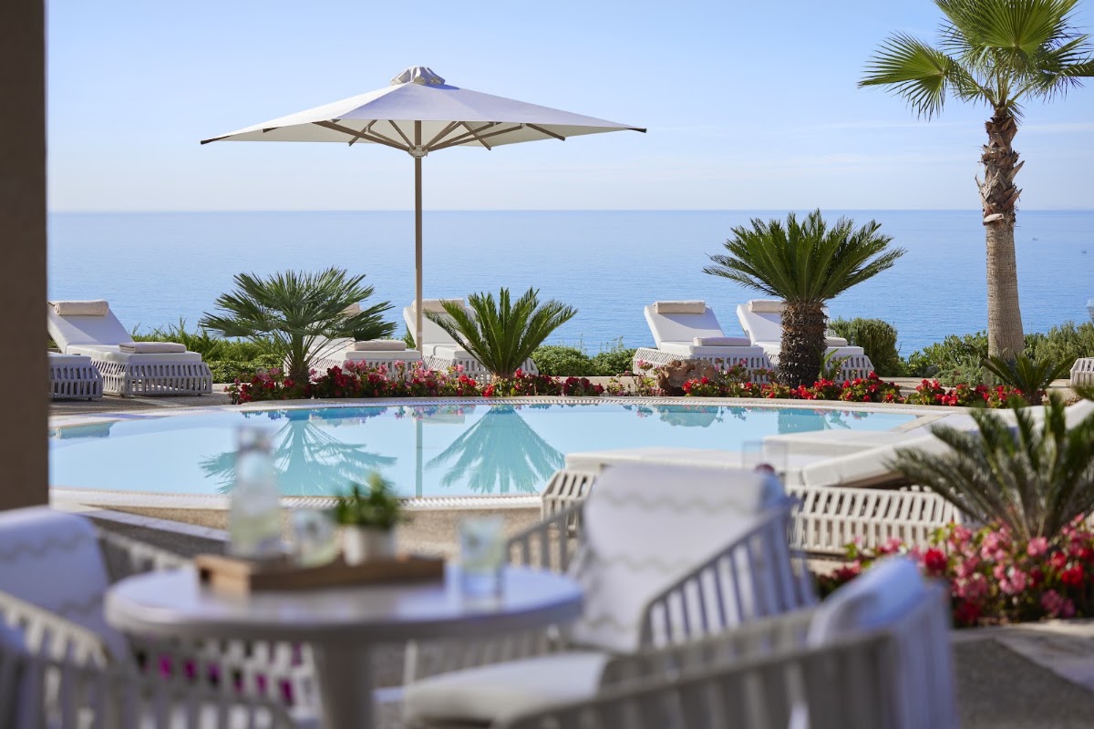 Pool area at Ikos Oceania with white umbrellas, loungers, palm trees, red flowers, and Mediterranean Sea beyond.