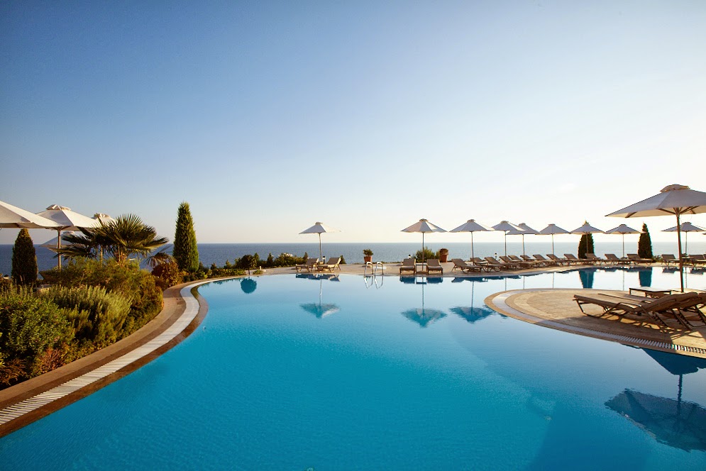 Curved infinity pool overlooking the Aegean Sea at Ikos Oceania, with loungers, umbrellas, and cypress trees along the deck under clear sky.