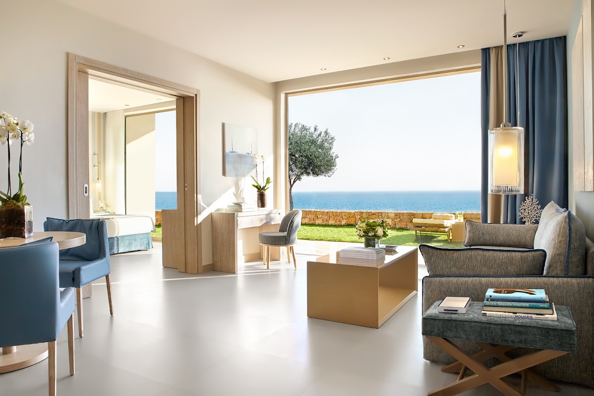 Modern suite interior at Ikos Oceania with sea view, featuring contemporary furniture, open layout, and access to terrace overlooking Mediterranean coastline.