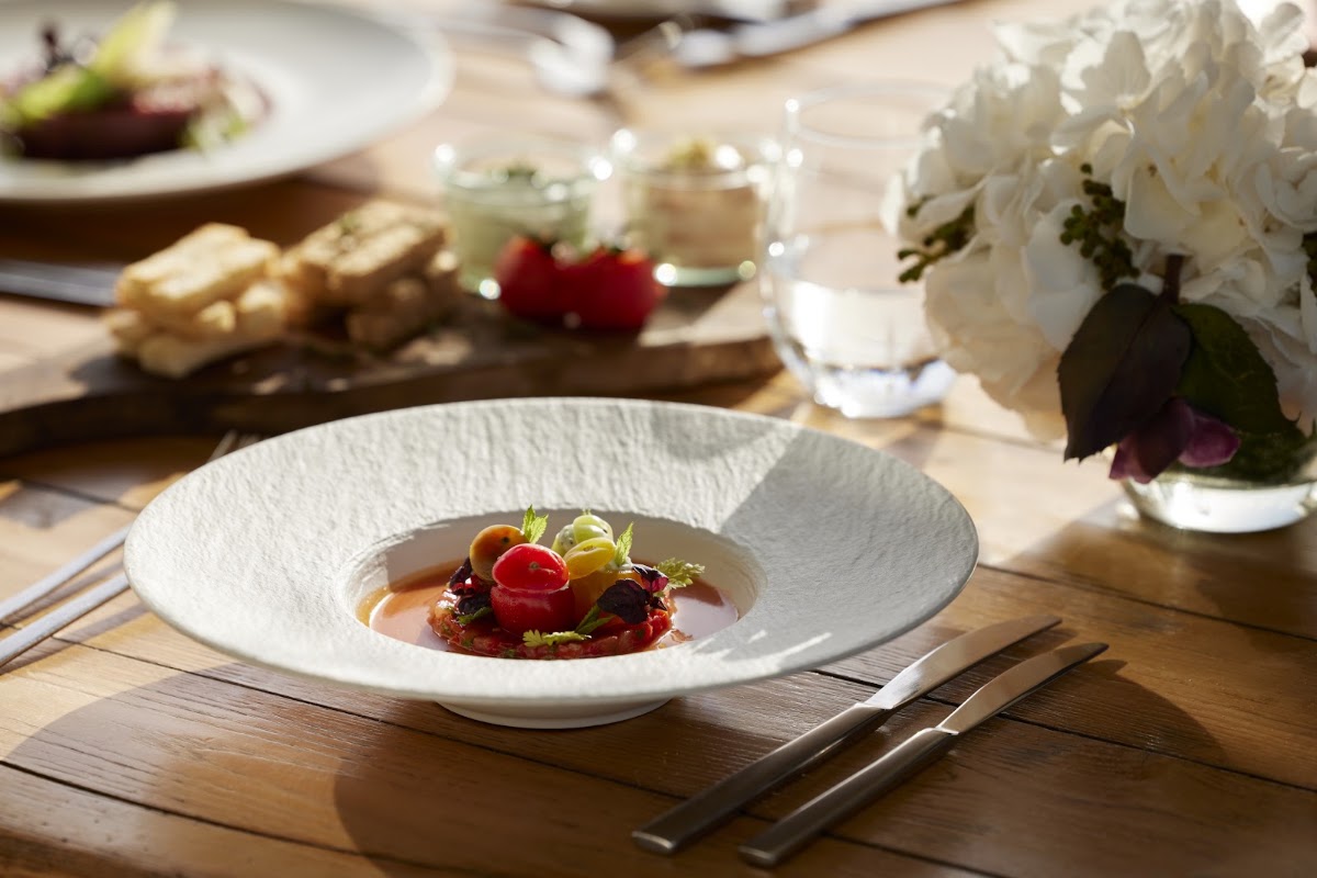 Fine dining plate with berries and sauce at wooden table, white flowers and place settings visible in background.