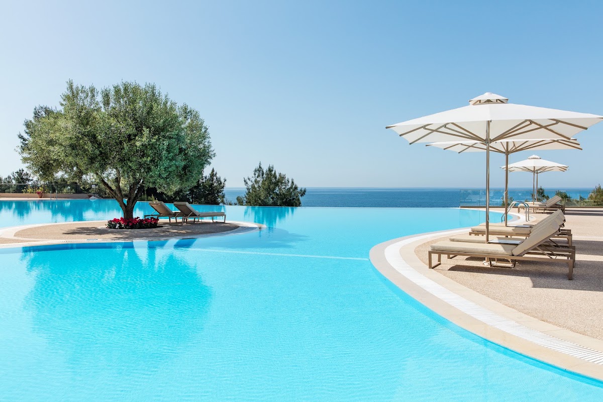 Large infinity pool with lounge chairs and cream umbrellas at Ikos Oceania, overlooking the sea with an olive tree on an island in the water.