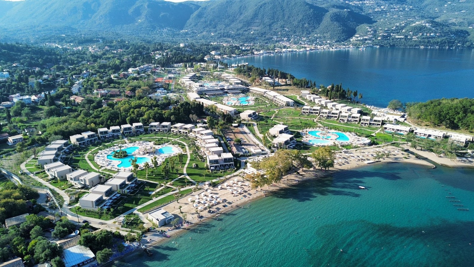 Aerial view of Ikos Odisha resort along a turquoise bay, with white buildings, multiple pools, beach with loungers, and forested mountains beyond.