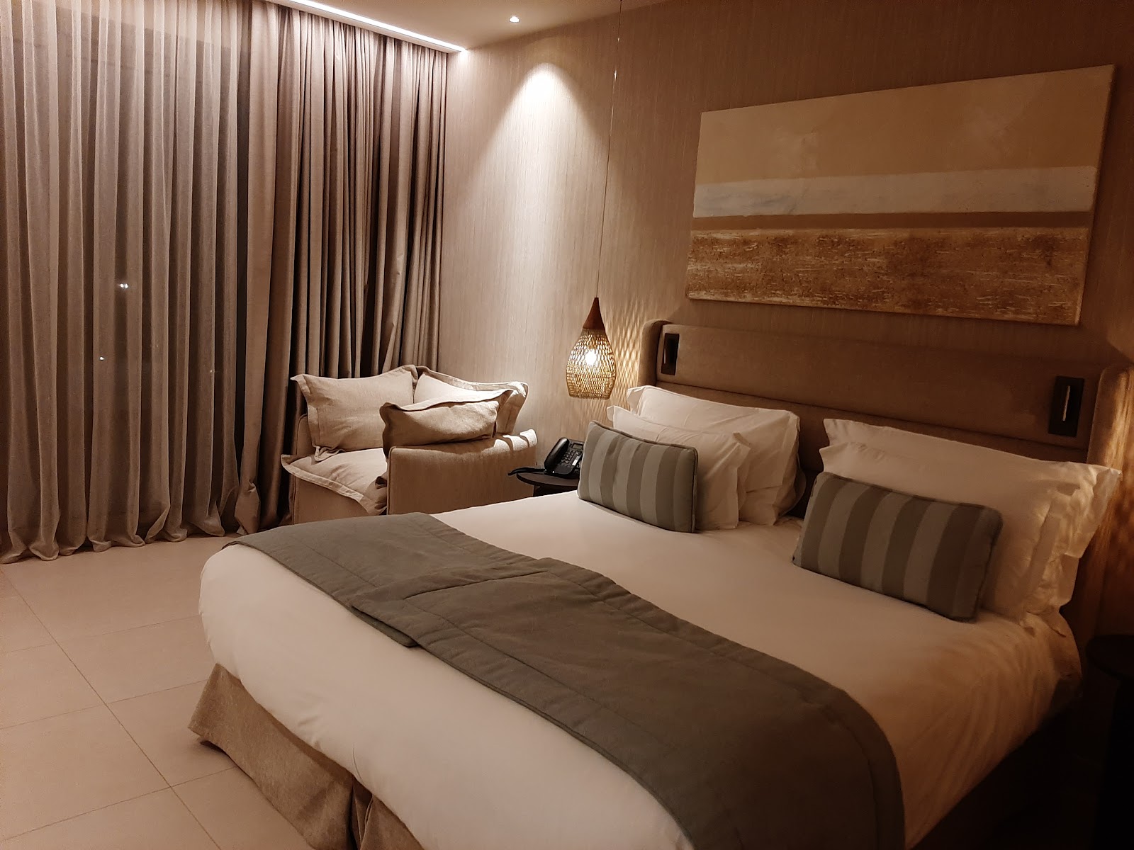 Resort guestroom with two beds dressed in cream linens and striped pillows, seating area, floor-to-ceiling curtains, and warm ambient lighting.