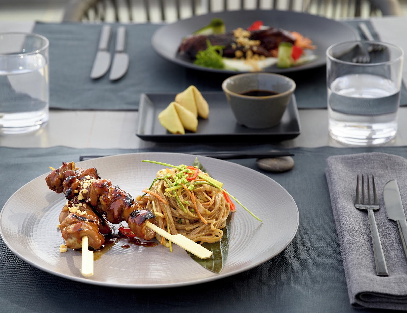 Plated Asian fusion dinner at Ikos Odisia with glazed chicken skewer and noodles, water glasses, and a sauce cup on gray table linens.