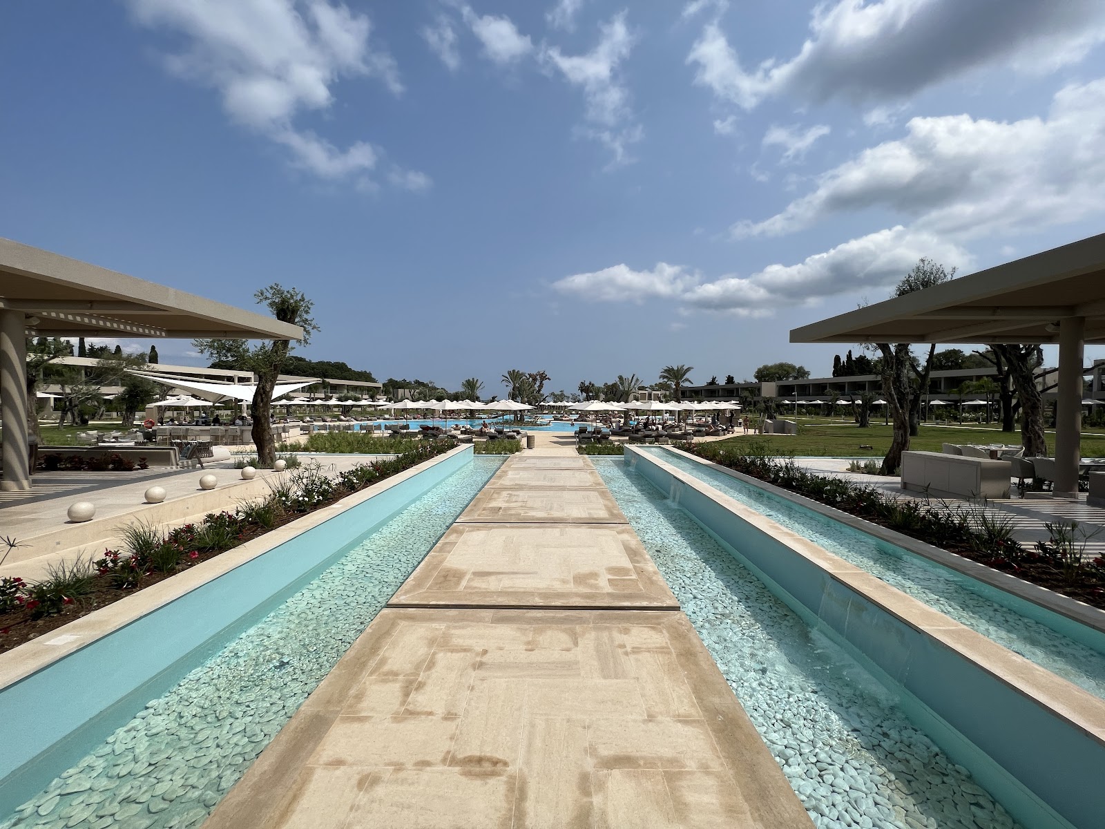 Narrow lap pools with turquoise water flank a stone walkway leading toward main pool area and shaded loungers at Ikos Odisia.