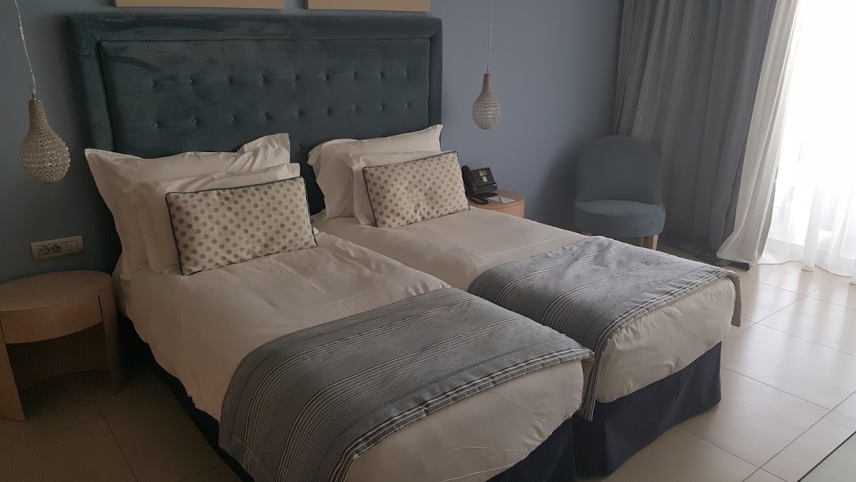 Two twin beds with tufted green headboard, cream linens, patterned pillows, and gray striped throws in a bright resort room with pendant lights.