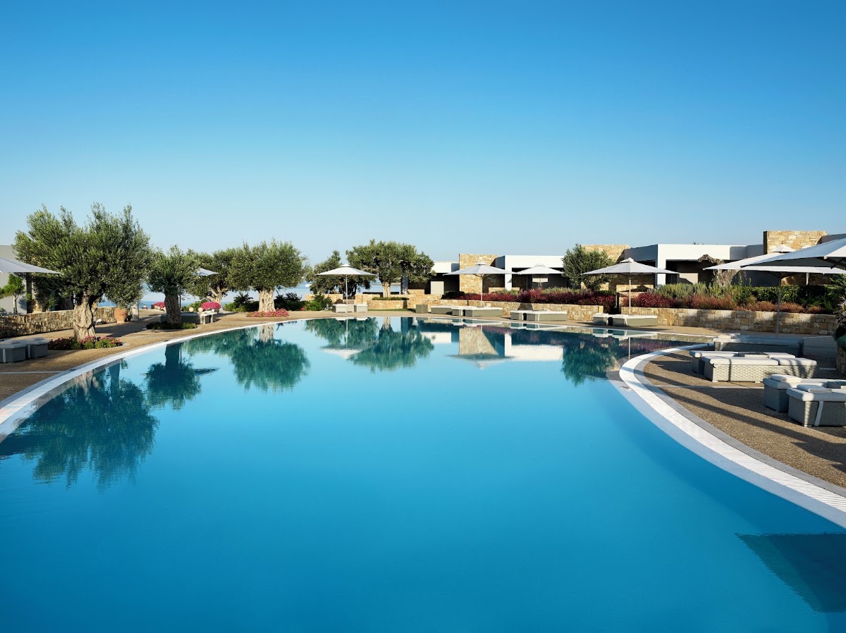 Large turquoise pool at Ikos Olivia with loungers, olive trees, and modern white buildings under clear blue sky.