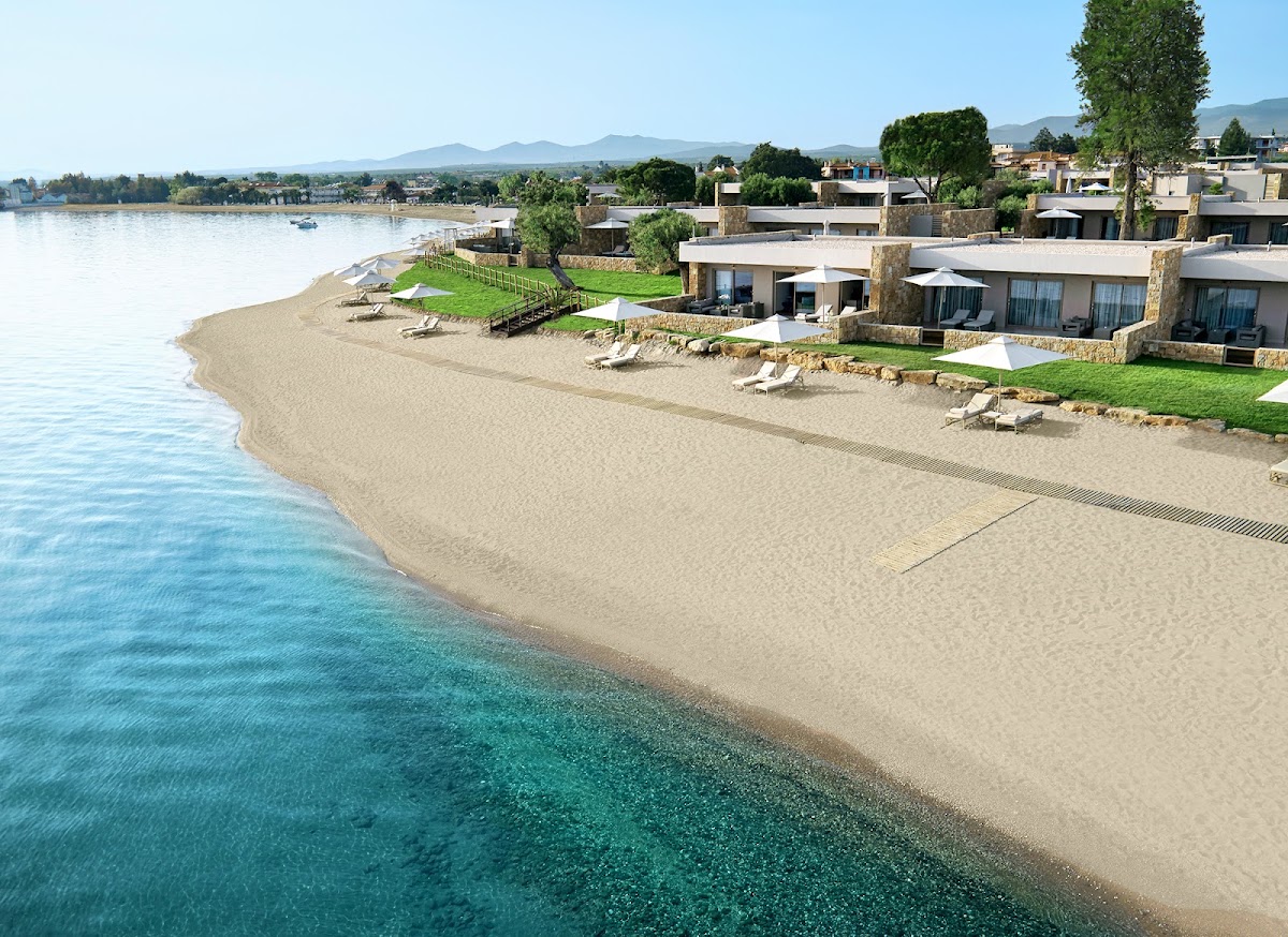 Beachfront villas with stone facades line a sandy beach at Ikos Olivia, with lounge chairs, umbrellas, and turquoise water below.