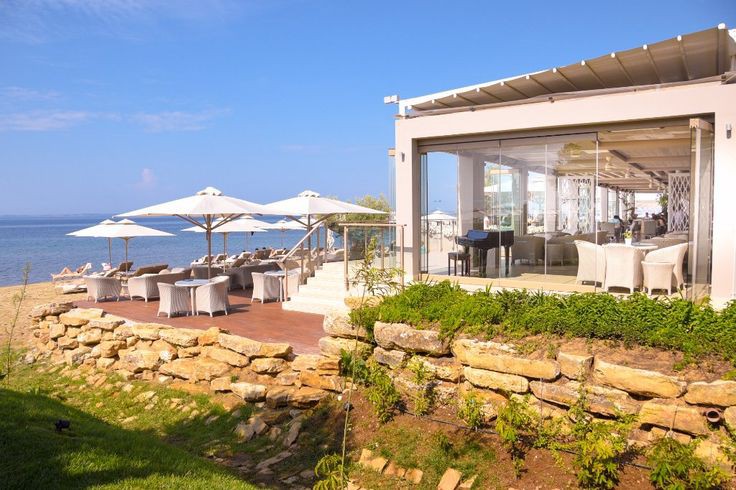 Beachfront terrace at Ikos Olivia with white umbrellas, loungers, and a modern pavilion overlooking the Aegean Sea.
