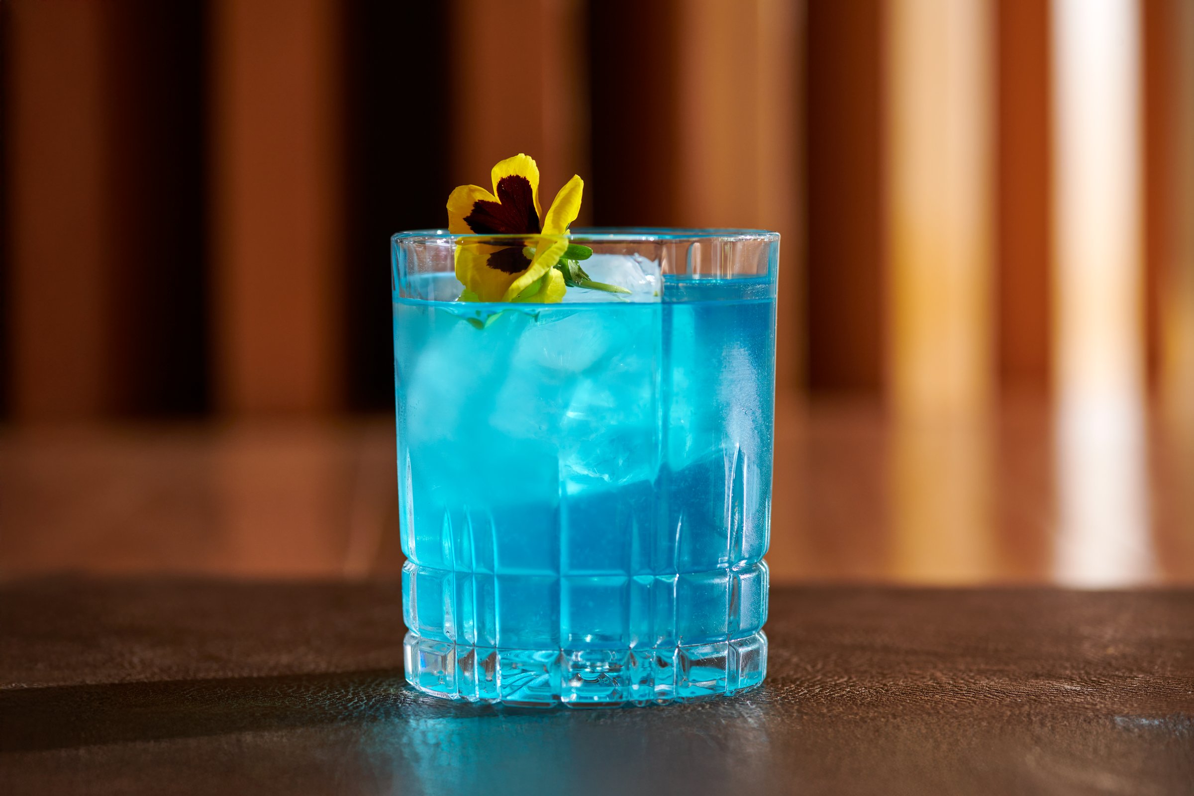 Bright blue cocktail in a textured glass garnished with a yellow and purple flower.