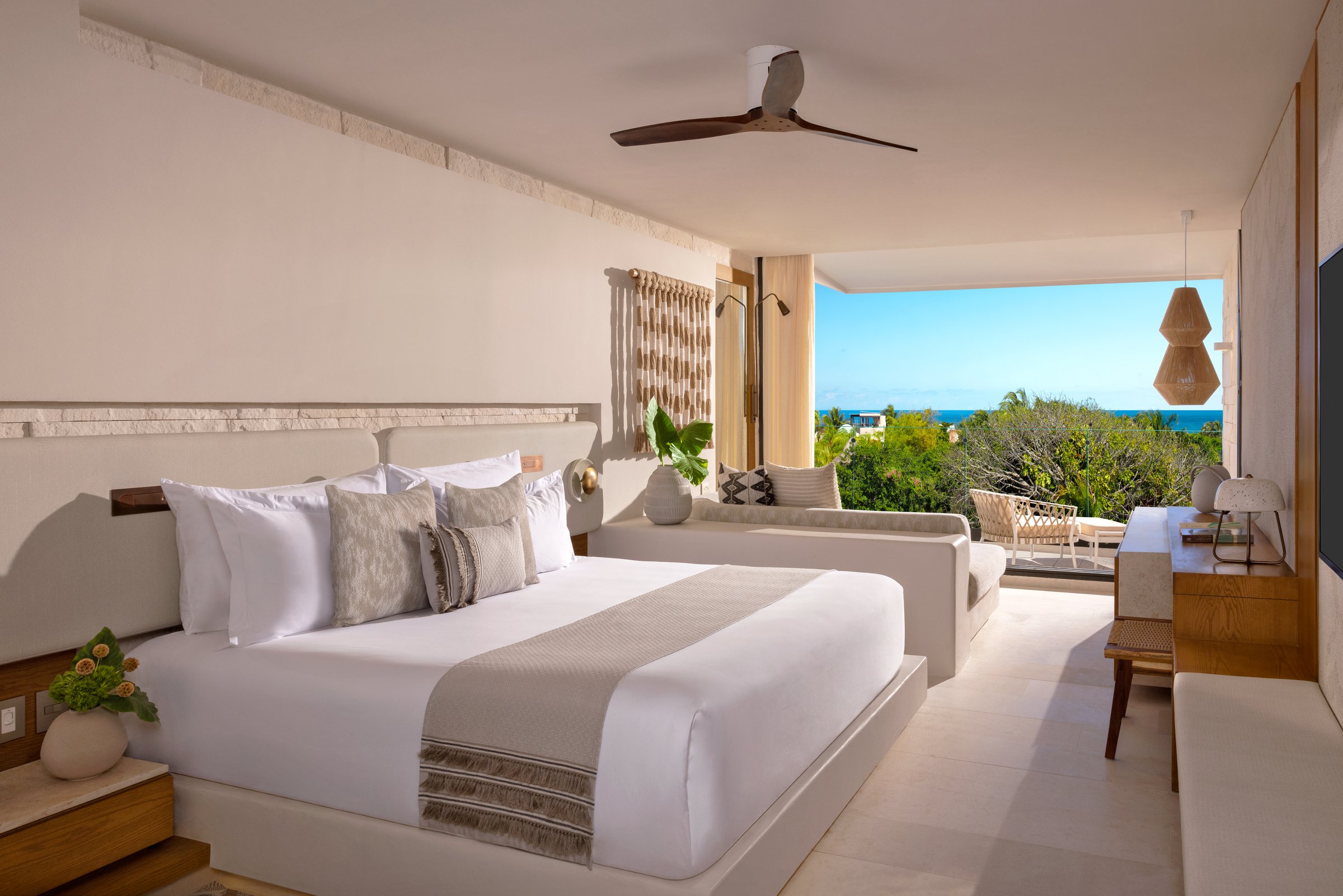 Junior suite with a king bed, light wood headboard and sliding doors to a balcony over green trees and the ocean.