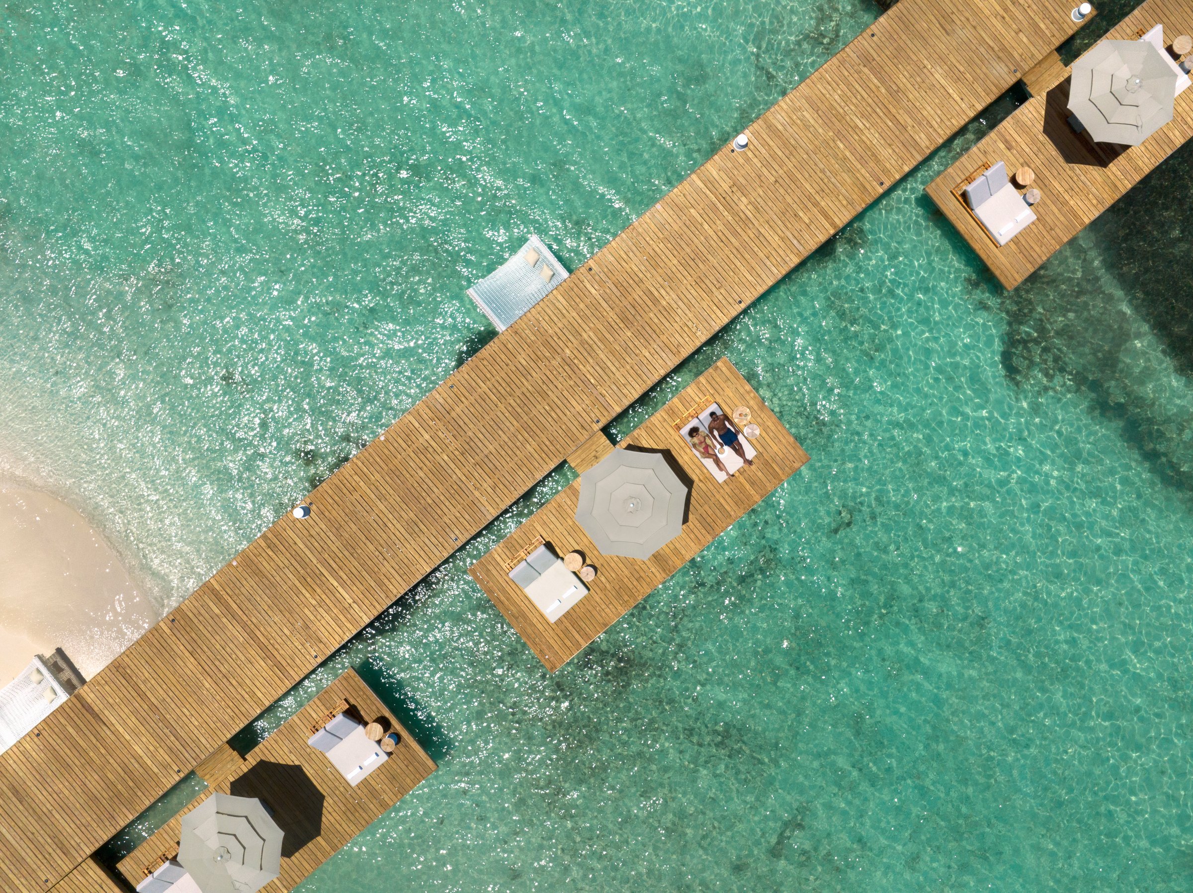 Top-down aerial of a wooden pier with square platforms holding white loungers and umbrellas over turquoise water.
