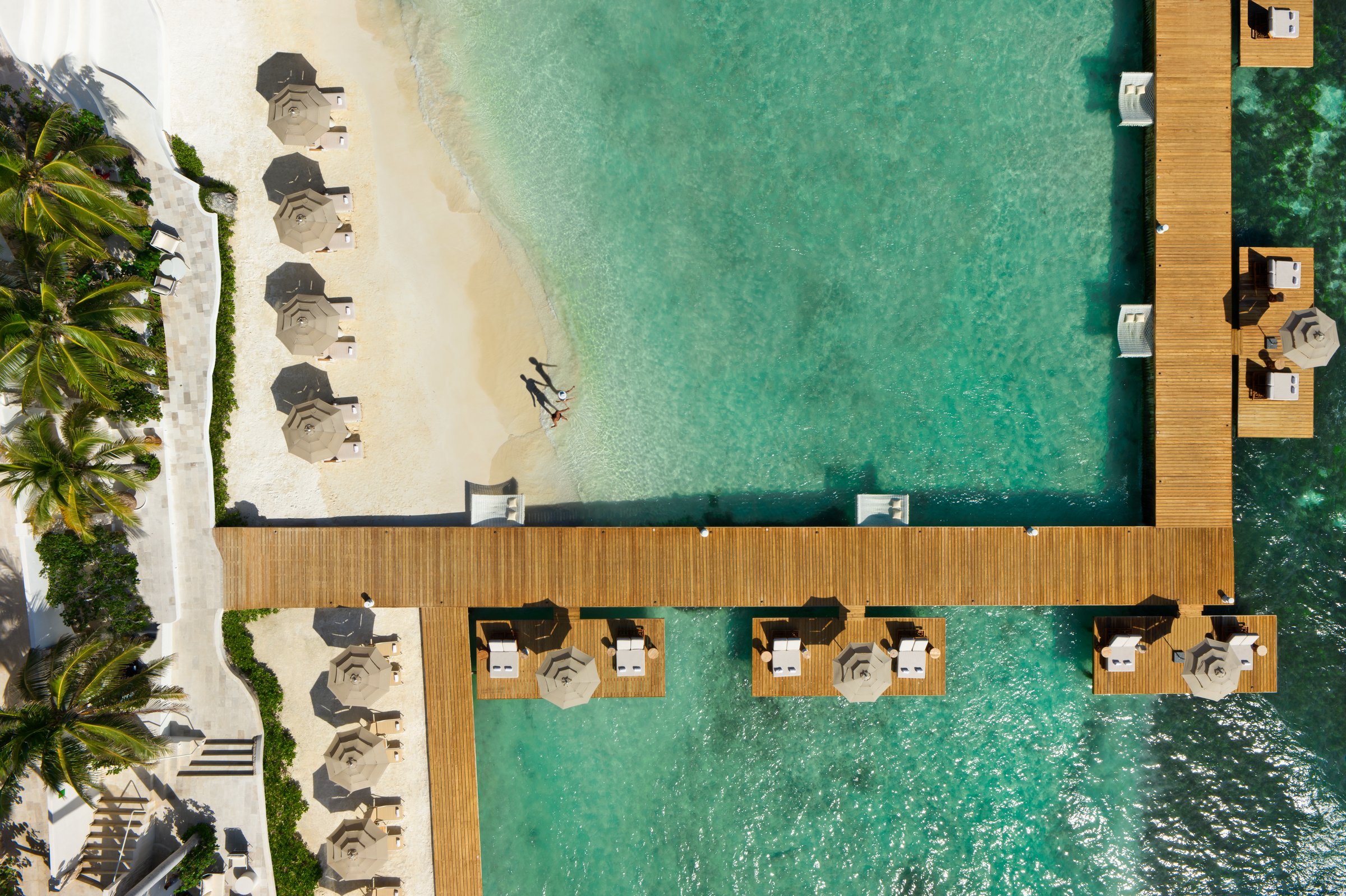Top-down aerial of the white sand beach with umbrellas, palms and a wooden pier over shallow turquoise water.