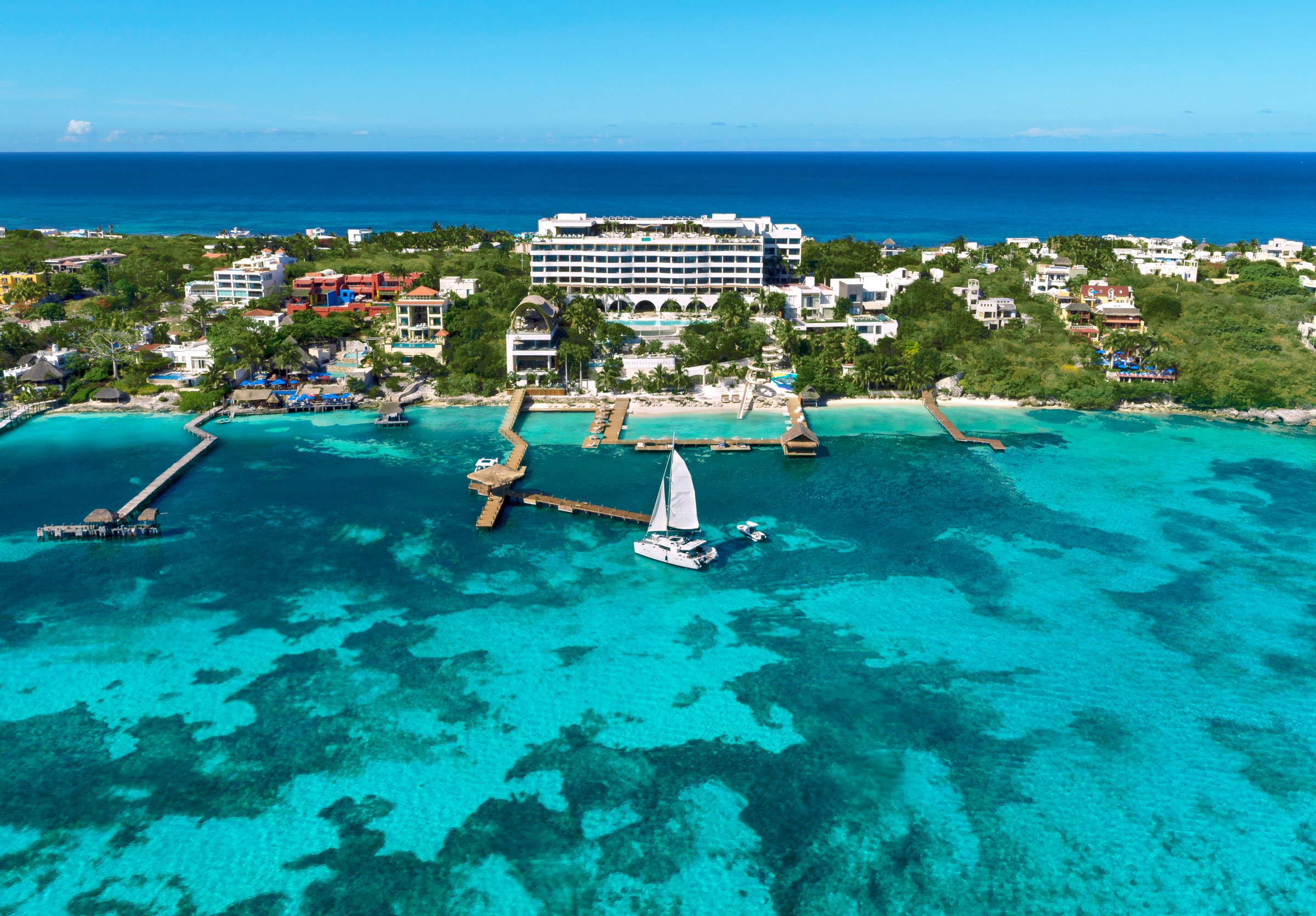 Aerial view of the white resort buildings among green foliage, with a long pier and sailboat on clear blue water.