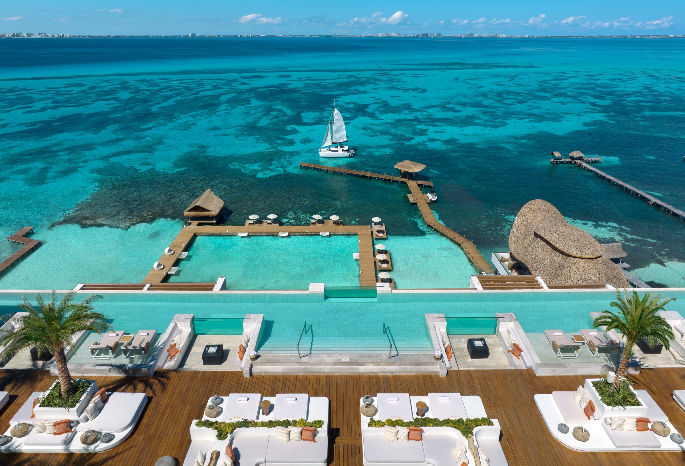 High-angle aerial of the rooftop infinity pool with white loungers and cabanas above the turquoise sea and a sailing catamaran.