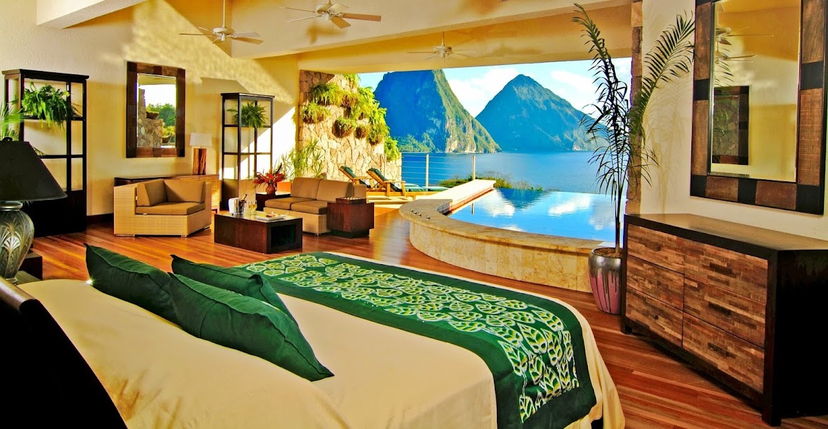 Luxury resort bedroom with cream bedding and green runner overlooking an infinity pool, lounge seating, and dramatic Piton mountains across the bay.