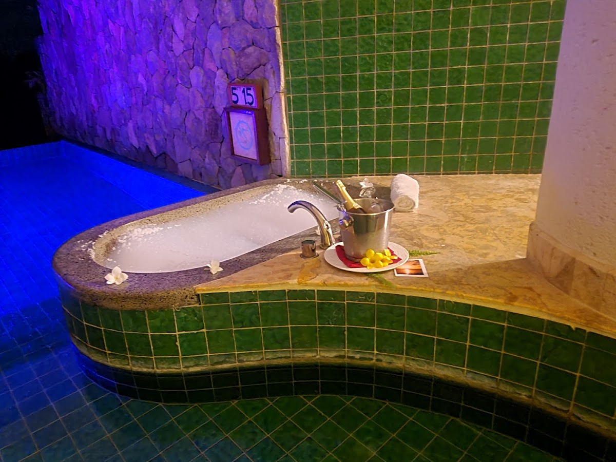 Curved stone bathtub with brass faucet beside a champagne bucket and fruit platter, surrounded by green tile walls and blue lighting at Jade Mountain.