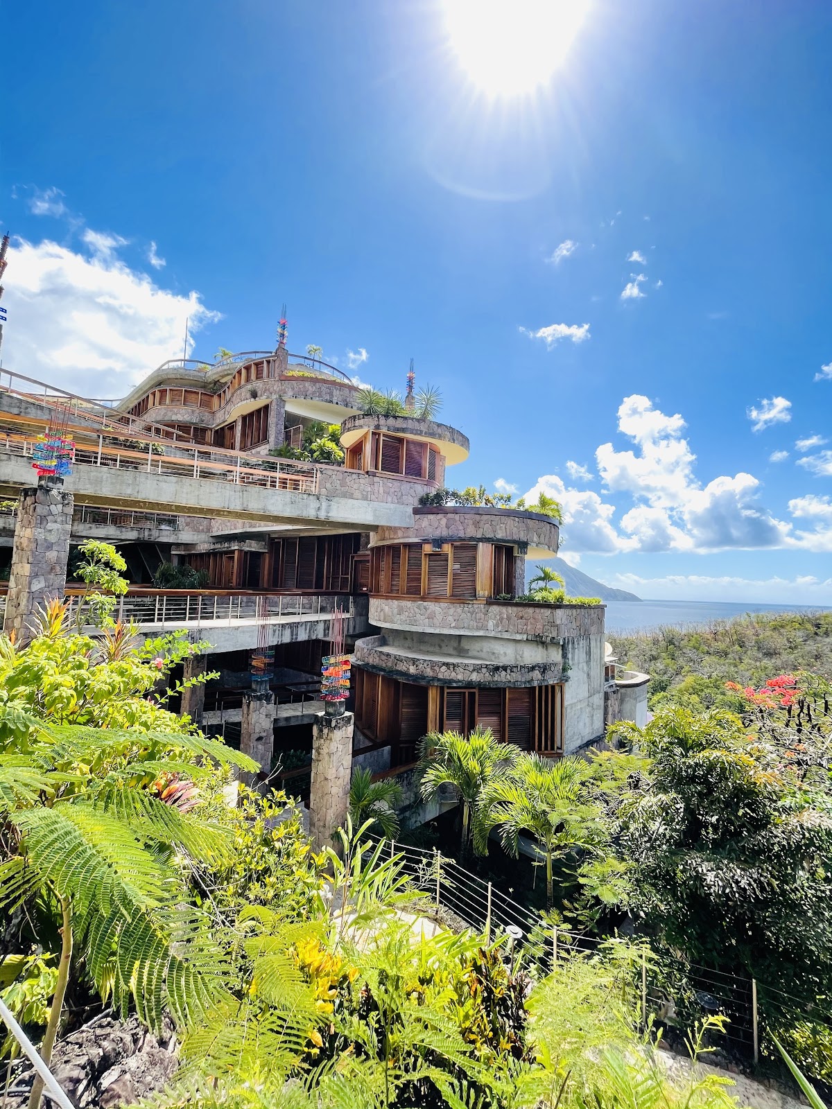 Multi-level concrete resort building with curved architecture and wooden shutters nestled in lush tropical vegetation, overlooking the Caribbean Sea and a distant mountain.