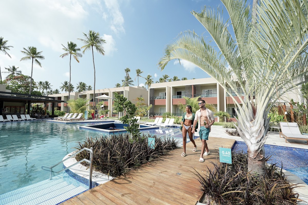Couple walks past a wooden deck with palm fronds overhead beside a turquoise pool at Japaratinga Lounge Resort, with modern buildings and tropical palms beyond.