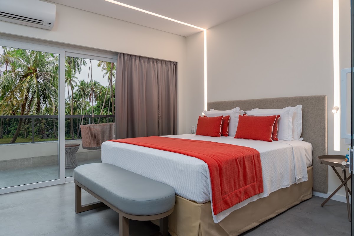 Modern resort bedroom with red accents, grey upholstered bed, and palm tree views through sliding glass doors at Japaratinga Lounge Resort.