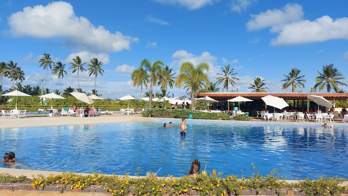 Guests swim in a large pool at Japaratinga Lounge Resort with palm trees, white umbrellas, and thatched-roof structures lining the sandy beach area under blue sky.