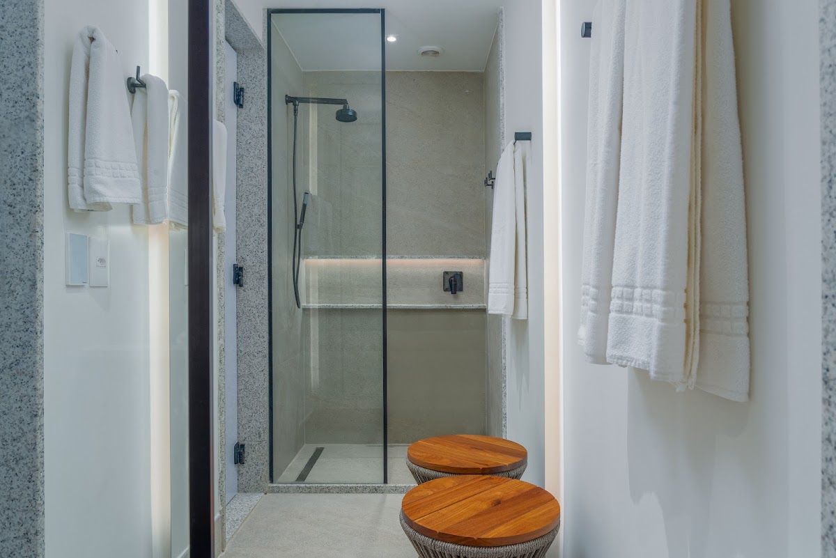 Modern bathroom with walk-in shower enclosed by glass door, terrazzo walls, white towel racks, and two round wooden stools in foreground.