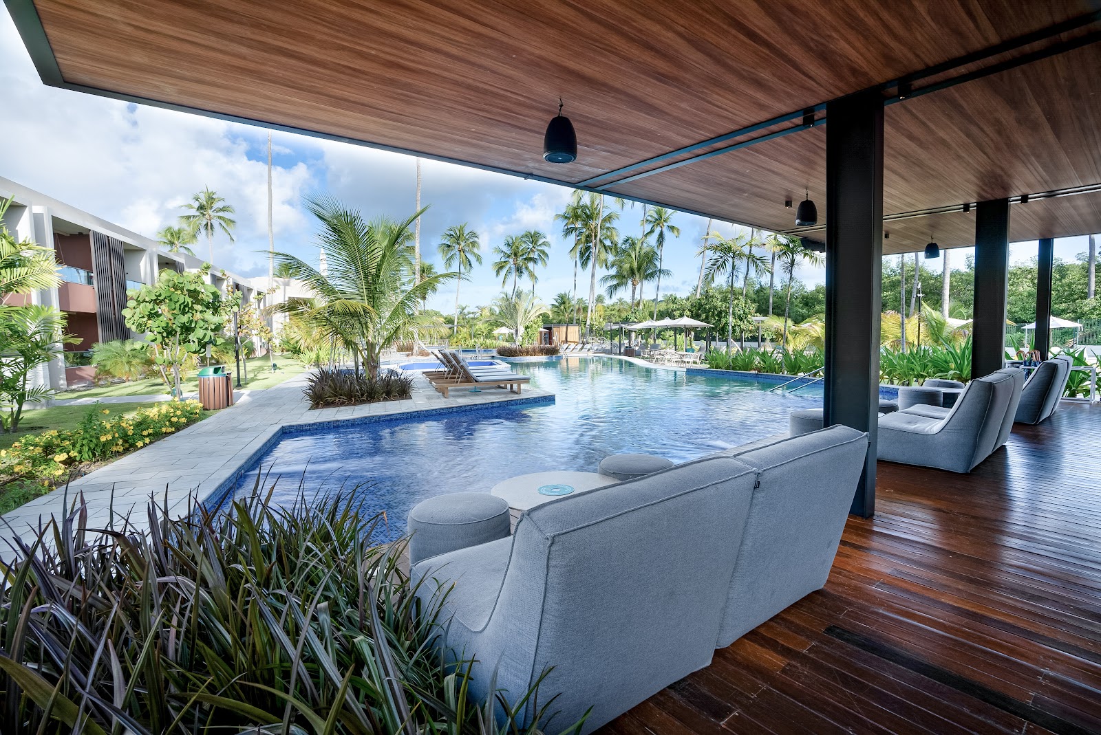 Large curved pool with swimmers, white loungers, pink multi-story hotel building, and coconut palms at Japaratinga Lounge Resort.
