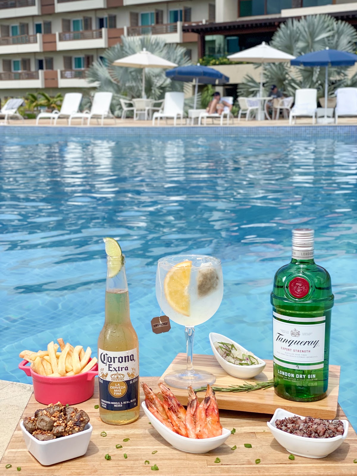Poolside spread at Japaratinga Lounge Resort featuring cocktails, beer, shrimp, fries, and appetizers on wooden deck with loungers and umbrellas behind.