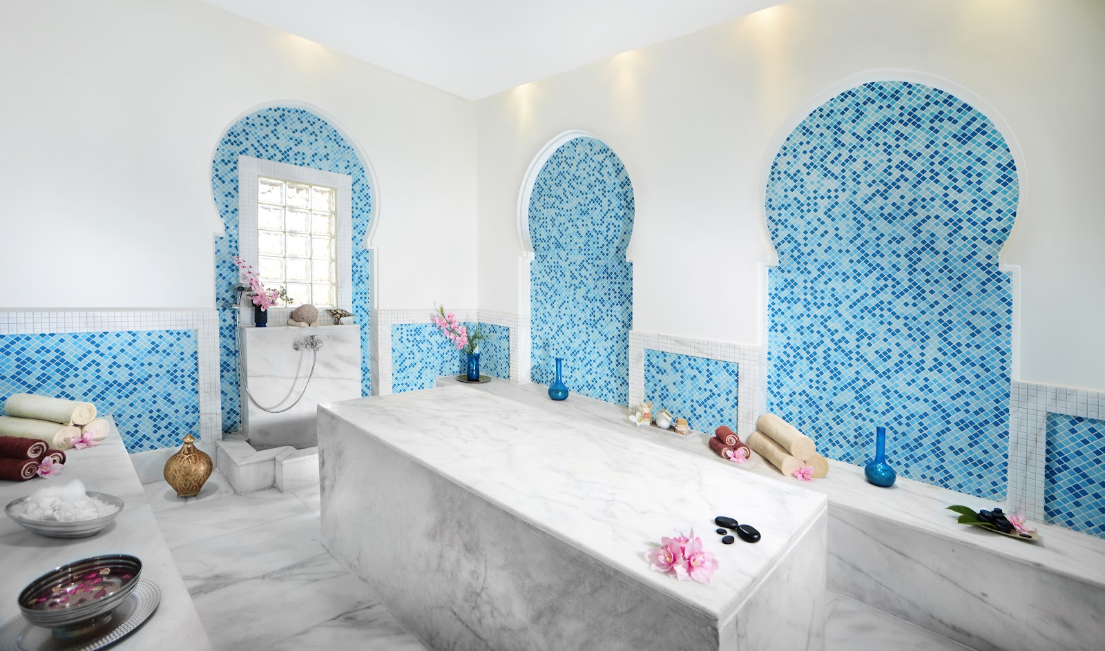 Spa bathroom with marble soaking tub, blue mosaic tile alcoves, pink flowers, and towels at Jaz Aquamarine Resort.